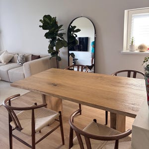 Peut inclure: Une table à manger en bois clair avec une finition naturelle, soutenue par un piédestal central. Quatre chaises avec des assises en osier et des dossiers en bois incurvés entourent la table. Un grand miroir en arche et une plante en pot sont visibles en arrière-plan.