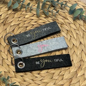 Könnte beinhalten: Drei rechteckige Schlüsselanhänger in Grau- und Schwarztönen, jeweils mit einem silbernen Schlüsselring. Die Schlüsselanhänger zeigen die Worte "BE you TIFUL" in goldener und rosa Schrift. Die Schlüsselanhänger liegen auf einer hellbraunen, gewebten Oberfläche.