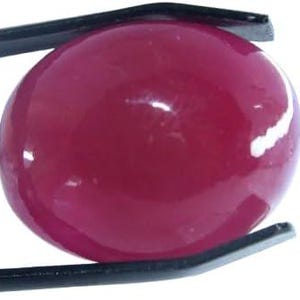 May include: A polished, oval-shaped, deep red gemstone held by black tweezers. The gem has a glossy surface, reflecting light. The tweezers are positioned to display the gem against a white background, highlighting its color and shape.