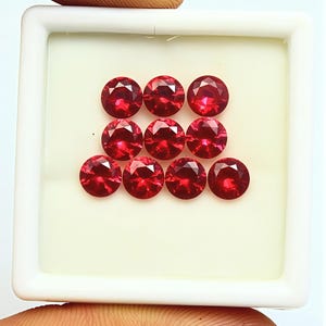 Puede incluir: Una colección de nueve piedras preciosas redondas, facetadas y de color rojo intenso, dispuestas en un patrón de cuadrícula dentro de una caja de exhibición cuadrada blanca. Cada piedra presenta un corte brillante con facetas.