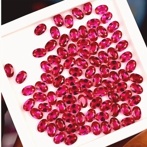 May include: A collection of numerous oval-cut, vibrant pinkish-red gemstones arranged within a square white display tray. Each faceted stone exhibits a brilliant, reflective surface, showcasing a consistent deep magenta hue across the entire set.
