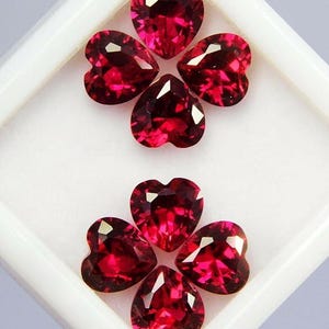 May include: Six heart-shaped, faceted, deep red gemstones arranged in two clover-like patterns. The gems are set against a white background within a white square container, showcasing their brilliant cut and color.