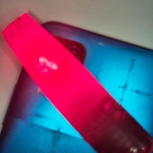Può includere: Un oggetto rettangolare rosa brillante con una superficie liscia e solida è posizionato diagonalmente sopra uno smartphone blu scuro. Lo smartphone presenta un ritaglio visibile per l'obiettivo della fotocamera e un pannello posteriore testurizzato.
