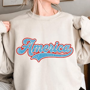 May include: A beige crewneck sweatshirt featuring the word America in a large, retro-style cursive font. The lettering is light blue with a distressed texture and is outlined in a bold red border, centred on the chest of the garment.