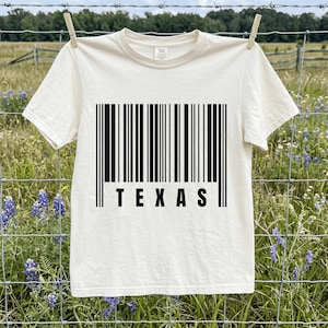 Peut inclure: Un t-shirt à manches courtes de couleur crème avec une impression graphique noire représentant un code-barres au centre de la poitrine. Sous le code-barres, le mot TEXAS est imprimé dans une police noire, grasse et sans empattement.