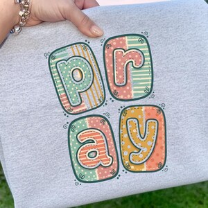 Peut inclure: Un sweat-shirt gris clair avec le mot "PRAY" en lettres colorées de style patchwork. Chaque lettre est soulignée en vert foncé et présente un motif unique, notamment des pois et des rayures. Le sweat-shirt est tenu par une personne.