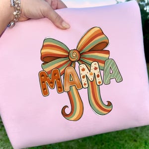 Peut inclure: Un coussin rose clair avec un nœud coloré et le mot "MAMA" aux couleurs d'automne. Le nœud présente des rayures orange, marron et vertes. Les lettres sont décorées de feuilles d'automne.