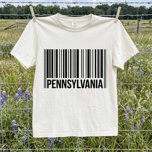 Könnte beinhalten: Ein cremefarbenes Kurzarm-T-Shirt mit einer schwarzen Strichcode-Grafik, die mittig auf der Brust aufgedruckt ist. Unter den vertikalen schwarzen Linien des Strichcodes steht das Wort PENNSYLVANIA in einer fetten, schwarzen serifenlosen Schrift.
