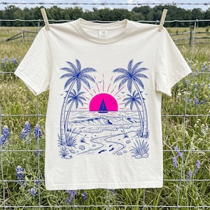 Peut inclure: Un t-shirt à manches courtes de couleur crème avec une impression graphique bleue et rose vif représentant une scène de plage. Le motif comprend des palmiers, un voilier sur l'océan, un soleil rose vif, des vagues et des empreintes de pas.