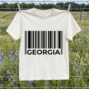 Peut inclure: Un t-shirt à manches courtes de couleur crème avec une impression graphique noire représentant un code-barres et le mot GEORGIA écrit en lettres capitales noires et grasses juste en dessous des lignes du code-barres sur le devant.