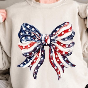 Peut inclure: Un sweat-shirt beige orné d'un grand nœud décoratif rouge, blanc et bleu, rappelant le drapeau américain. Le nœud présente des rayures et des étoiles, créant un thème patriotique. Le sweat-shirt a des manches longues.