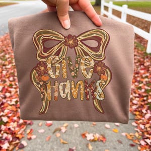Peut inclure: Un sweat-shirt marron avec un nœud décoratif et l'inscription "GIVE THANKS" dans un design sur le thème de l'automne. Le texte et le nœud présentent des accents floraux et de citrouille, parfaits pour la saison de Thanksgiving.