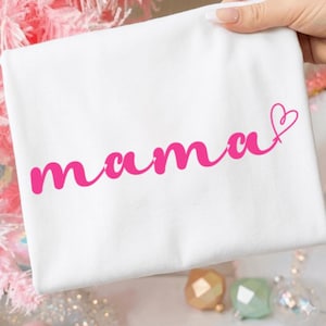 Peut inclure: T-shirt blanc avec le mot "mama" en rose écrit en cursive. Un cœur rose est dessiné à la fin du mot. Le t-shirt est tenu par une main.