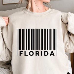 Peut inclure: Un sweat-shirt à col rond beige avec une impression graphique noire au centre de la poitrine. Le motif se compose d'un code-barres vertical avec le mot FLORIDA imprimé en lettres capitales noires, grasses et sans empattement en dessous.