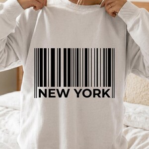 Peut inclure: Un sweat-shirt à col rond gris clair avec une impression graphique noire représentant un code-barres centré sur la poitrine. Sous le code-barres, le texte NEW YORK est imprimé dans une police noire, grasse et sans empattement. Manches longues.