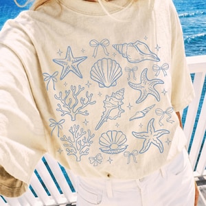 Peut inclure: Un t-shirt couleur crème arborant un imprimé graphique bleu clair représentant divers coquillages, étoiles de mer, branches de corail et petits nœuds dispersés sur le devant. Le motif inclut plusieurs étoiles à quatre branches entre les éléments.