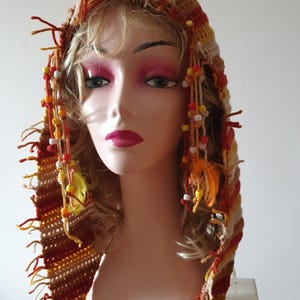May include: A handmade crochet hood in shades of orange, brown, and cream, adorned with dangling strands of yarn, beads, and small feathers. The hood is displayed on a mannequin head, showcasing its intricate design and texture.