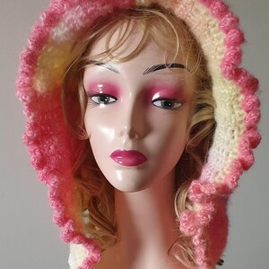 May include: A handmade, pink and white crochet hood with a ruffled edge. The hood is displayed on a mannequin head with blonde hair and pink makeup. The hood is a soft, textured accessory.