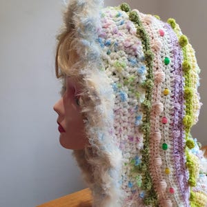 May include: A handmade crochet hat in pastel shades, with a fluffy, cream-coloured trim. The hat is decorated with green, pink, and yellow bobbles and a decorative green border. The hat is displayed on a mannequin head.