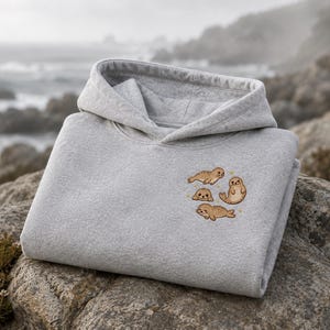 May include: A light gray hooded sweatshirt featuring an embroidered design on the chest. The embroidery depicts four small, brown, cartoon-style seals in various poses, accompanied by tiny yellow star accents scattered around the seal figures.