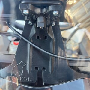 May include: Black plastic bracket with the text "WACKOM" and a bear logo. The bracket has a series of holes and mounting points, and a black cable is visible. The bracket is attached to a larger object.