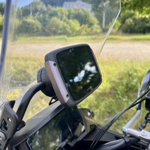Könnte beinhalten: Ein Motorrad-GPS-Gerät, das am Lenker montiert ist. Das Gerät hat einen schwarzen Bildschirm mit einem silbernen und schwarzen Rahmen. Die Windschutzscheibe des Motorrads ist im Hintergrund zu sehen, zusammen mit einer grünen Wiese und Bäumen.