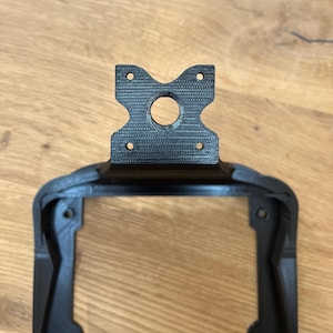 May include: A black, 3D-printed bracket with a distinctive shape. The top portion features a central hole and four smaller holes, while the lower section has a rectangular frame. The bracket is designed for mounting or support.