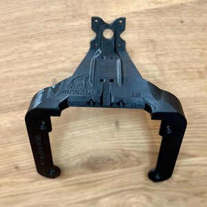 May include: A black, 3D-printed bracket with a unique shape, featuring the text "MACKOM" and "KV2". The bracket has a central mounting hole and two curved arms with additional holes. The design appears to be functional and industrial.