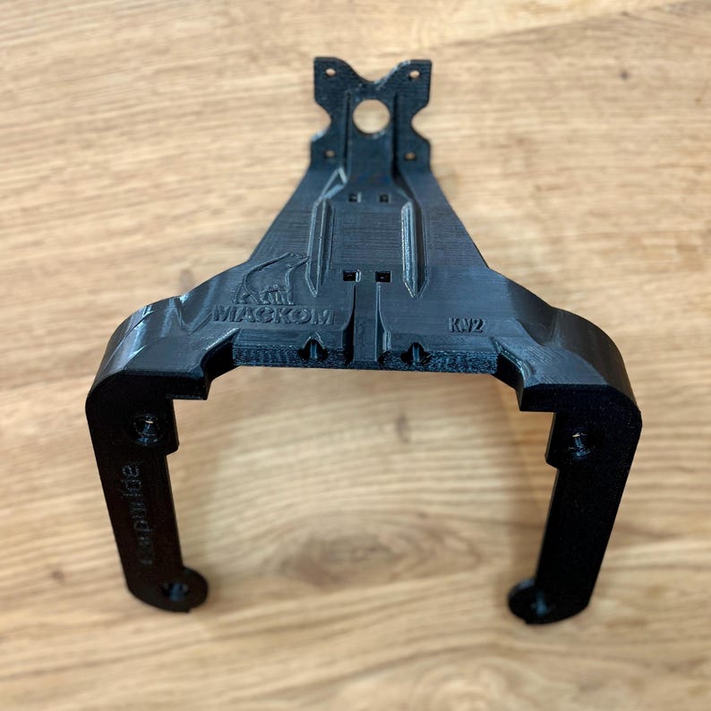 May include: A black, 3D-printed bracket with a unique shape, featuring the text "MACKOM" and "KV2". The bracket has a central mounting hole and two curved arms with additional holes. The design appears to be functional and industrial.