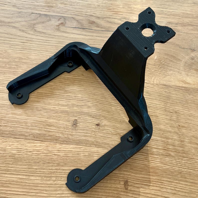 May include: A black, 3D-printed bracket with a unique, angular design. The bracket features multiple mounting holes and a central circular opening. It is made of a matte black material and sits on a wooden surface.