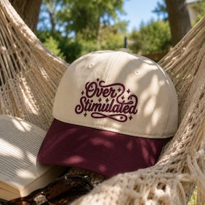 Peut inclure: Une casquette de baseball bicolore avec une calotte couleur crème et une visière bordeaux foncé. L'avant de la casquette présente le texte brodé "Over Stimulated" dans une police cursive bordeaux foncé, agrémenté de petites étoiles brodées.