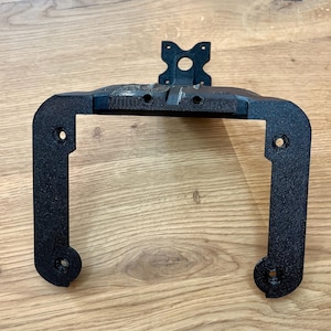 May include: A black, 3D-printed bracket with a rectangular frame and a mounting plate. The bracket has rounded corners and several holes for screws. The mounting plate has a circular hole and two smaller holes. The bracket is designed to hold an electronic device.