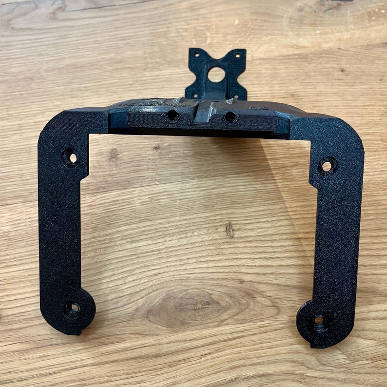 May include: A black, 3D-printed bracket with a rectangular frame and a mounting plate. The bracket has rounded corners and several holes for screws. The mounting plate has a circular hole and two smaller holes. The bracket is designed to hold an electronic device.