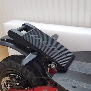 Op de afbeelding: Zwarte voetsteun voor een elektrische scooter met een getextureerd oppervlak en een handvat. De voetsteun is gemonteerd aan de achterkant van de scooter, bij het wiel. De scooter heeft een zwart spatbord en een rode vering.