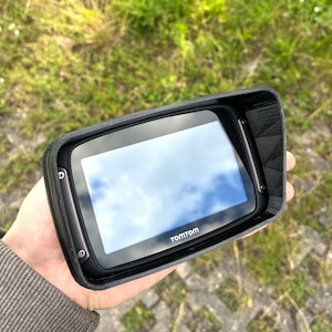 May include: A black TomTom GPS device with a protective visor. The screen reflects a cloudy sky. The device is held in a hand against a background of green grass.