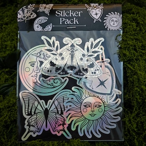 Könnte beinhalten: Ein Sticker-Pack mit holografischen Aufklebern mit himmlischen und von der Natur inspirierten Designs. Die Aufkleber zeigen eine Motte, einen Schmetterling, eine Sonne, einen Mond und ein Herz mit einem Schwert. Die Verpackung ist schwarz mit dem weißen Schriftzug "Sticker Pack".