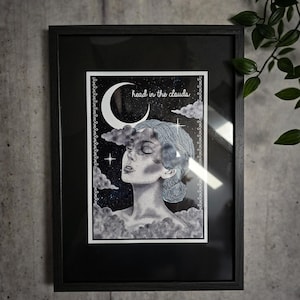 Könnte beinhalten: Schwarz gerahmter Kunstdruck mit einer Graustufen-Illustration einer Person mit geschlossenen Augen, umgeben von Wolken und einem Halbmond. Der Text "head in the clouds" befindet sich über dem Kopf der Person. Das Kunstwerk ist vor einem dunklen, sternenübersäten Hintergrund.