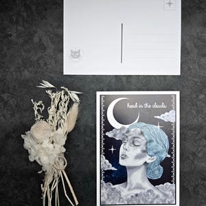 Könnte beinhalten: Eine Postkarte mit dem Text "head in the clouds" und einer Illustration eines Frauengesichts, umgeben von Wolken und Sternen. Eine leere Postkarte und ein kleiner Strauß Trockenblumen sind ebenfalls vorhanden.