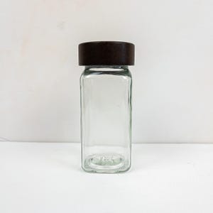 May include: A clear, rectangular glass jar with a dark brown lid. The jar is empty and has a simple, minimalist design. The lid is flat and covers the entire top of the jar. The jar is likely used for storing spices or other dry goods.