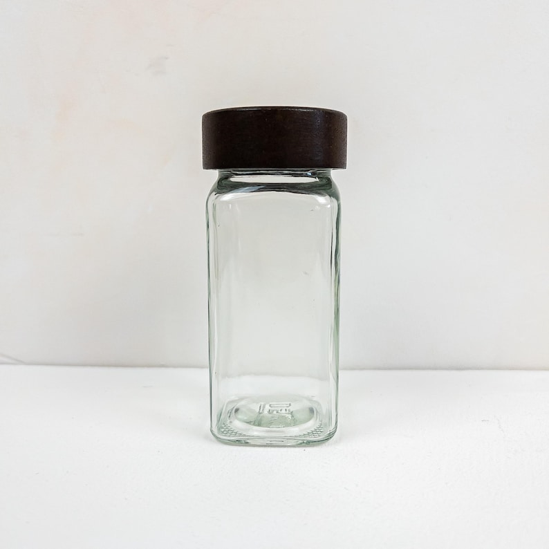 May include: A clear, rectangular glass jar with a dark brown lid. The jar is empty and has a simple, minimalist design. The lid is flat and covers the entire top of the jar. The jar is likely used for storing spices or other dry goods.