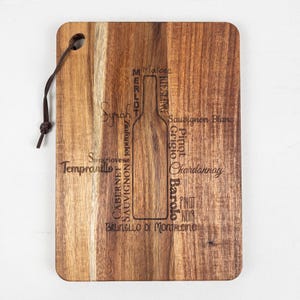 Op de afbeelding: Een rechthoekige houten snijplank met een donkerbruine leren lus. De plank heeft een omtrek van een wijnfles met ingegraveerde wijnsoorten, waaronder Syrah, Cabernet Sauvignon en Chardonnay.