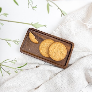 Può includere: Un vassoio rettangolare in legno scuro mostra tre biscotti rotondi marrone dorato, uno parzialmente mangiato. Il vassoio poggia su un panno bianco strutturato, con rametti verdi sullo sfondo. I biscotti sembrano essere fatti di avena o un cereale simile.