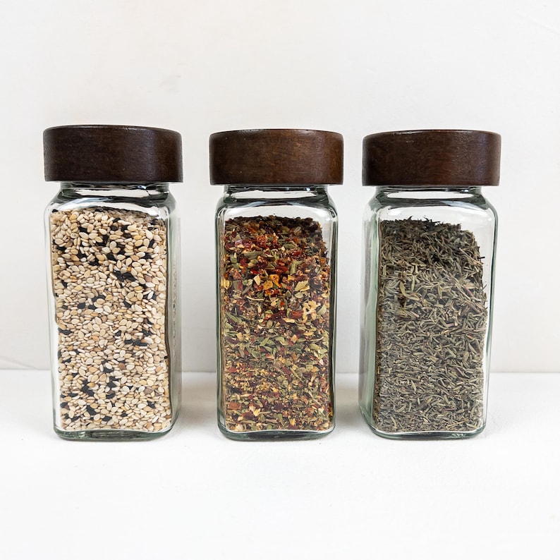 May include: Three glass spice jars with dark wooden lids. The jars are filled with different spices: sesame seeds, a red and green herb mix, and dried thyme. The jars are rectangular and sit on a white surface.