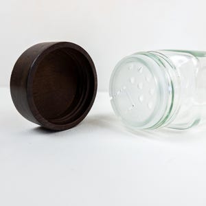 May include: A clear glass spice jar with a dark brown wooden lid. The jar has a shaker top with multiple small holes. The lid is separate from the jar, revealing the inside of the jar.