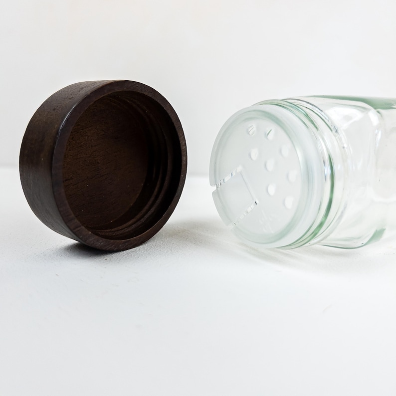 May include: A clear glass spice jar with a dark brown wooden lid. The jar has a shaker top with multiple small holes. The lid is separate from the jar, revealing the inside of the jar.