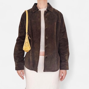 Könnte beinhalten: Braune Wildlederjacke mit Knopfleiste und Kragen. Eine kleine, gelbe Handtasche hängt über der Schulter. Die Jacke wird über einem weißen Oberteil und Rock getragen. Der Gesamtstil ist Vintage.