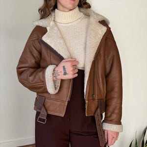 May include: A brown leather jacket with a cream shearling lining and collar. The jacket features a zip closure, a buckled belt at the waist, and shearling trim at the cuffs. The model is wearing a cream turtleneck sweater and brown trousers.
