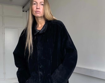 May include: A long, black coat with a button-up front and pockets. The coat has a velvet-like texture and a dark collar. The model is wearing the coat, and the background is a plain white wall.