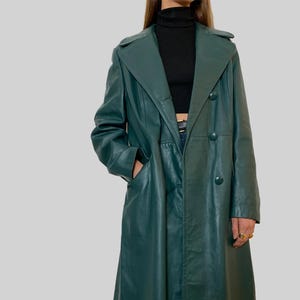 May include: A long, emerald green leather trench coat with a double-breasted design. The coat features a notched collar, pockets, and button closures. The coat is knee-length and has a classic, tailored silhouette.
