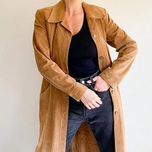 May include: A long, tan suede coat with a notched collar and button fastenings. The coat features vertical seam detailing and a tailored fit. The model is wearing a black top and dark wash jeans. The coat has a classic, vintage style.